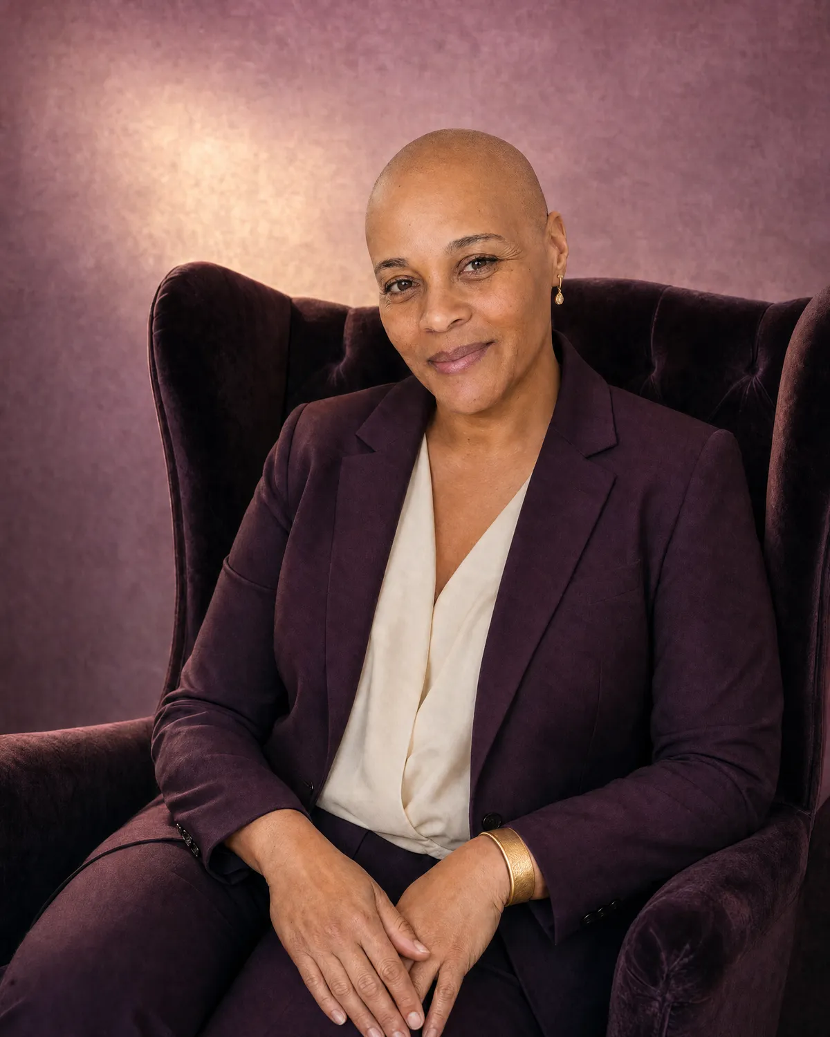 Jackie Allison in a deep plum suit with a gold cuff bracelet, seated upright in a plum velvet wingback chair against a warm plum backdrop.