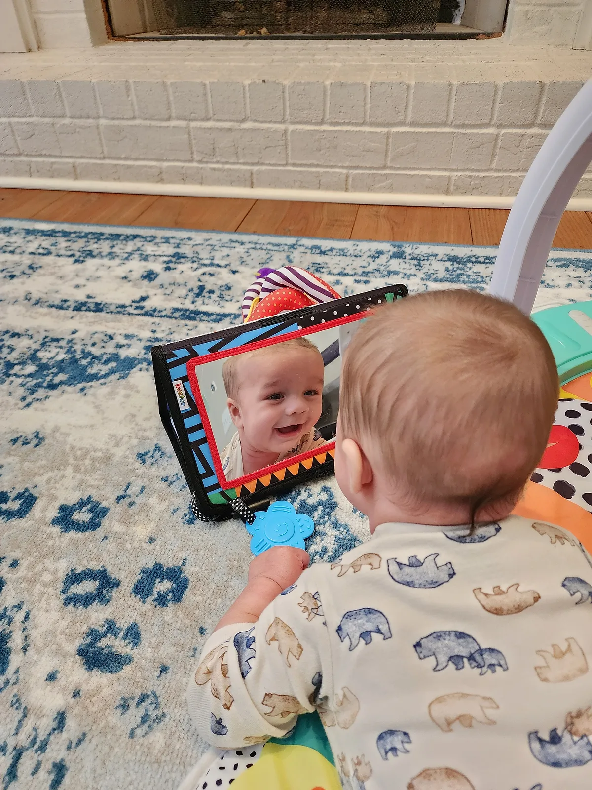 A baby lying on a play mat smiles at their reflection in a soft baby mirror.